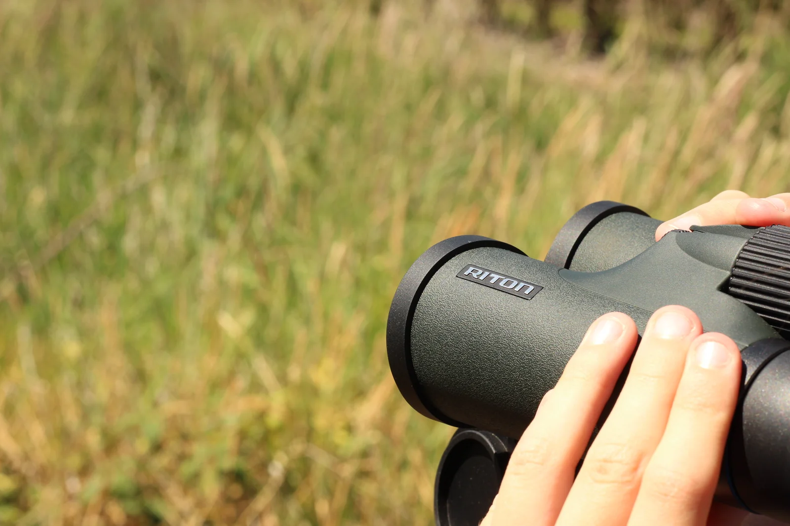 Tight close-up of the objective end of the Riton 5 Primal 10x42 ED held in the eleven-year-old's hand, the RITON wordmark prominent on the barrel, the objective bezel visible, and an out-of-focus grass background filling the frame