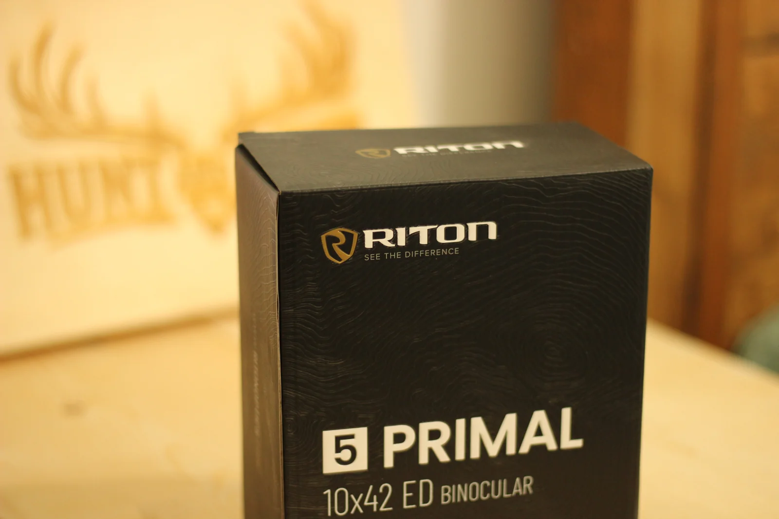 The Riton 5 Primal 10x42 ED retail box on a workbench, photographed with the Hunt and Live wood-burned antlers-and-deer-stencil logo on the board behind it, black topographic-pattern box with the white Riton shield and 5 PRIMAL 10x42 ED wordmark