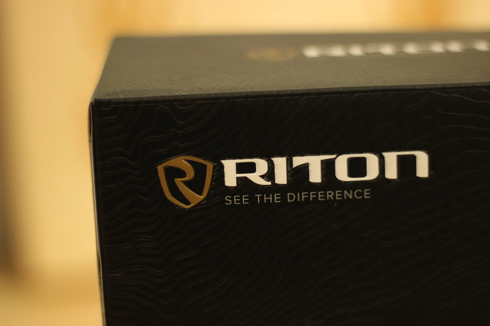 Close-up of the Riton shield logo and SEE THE DIFFERENCE wordmark on the upper corner of the 5 Primal 10x42 ED retail box, gold-and-white shield on black topographic-pattern background