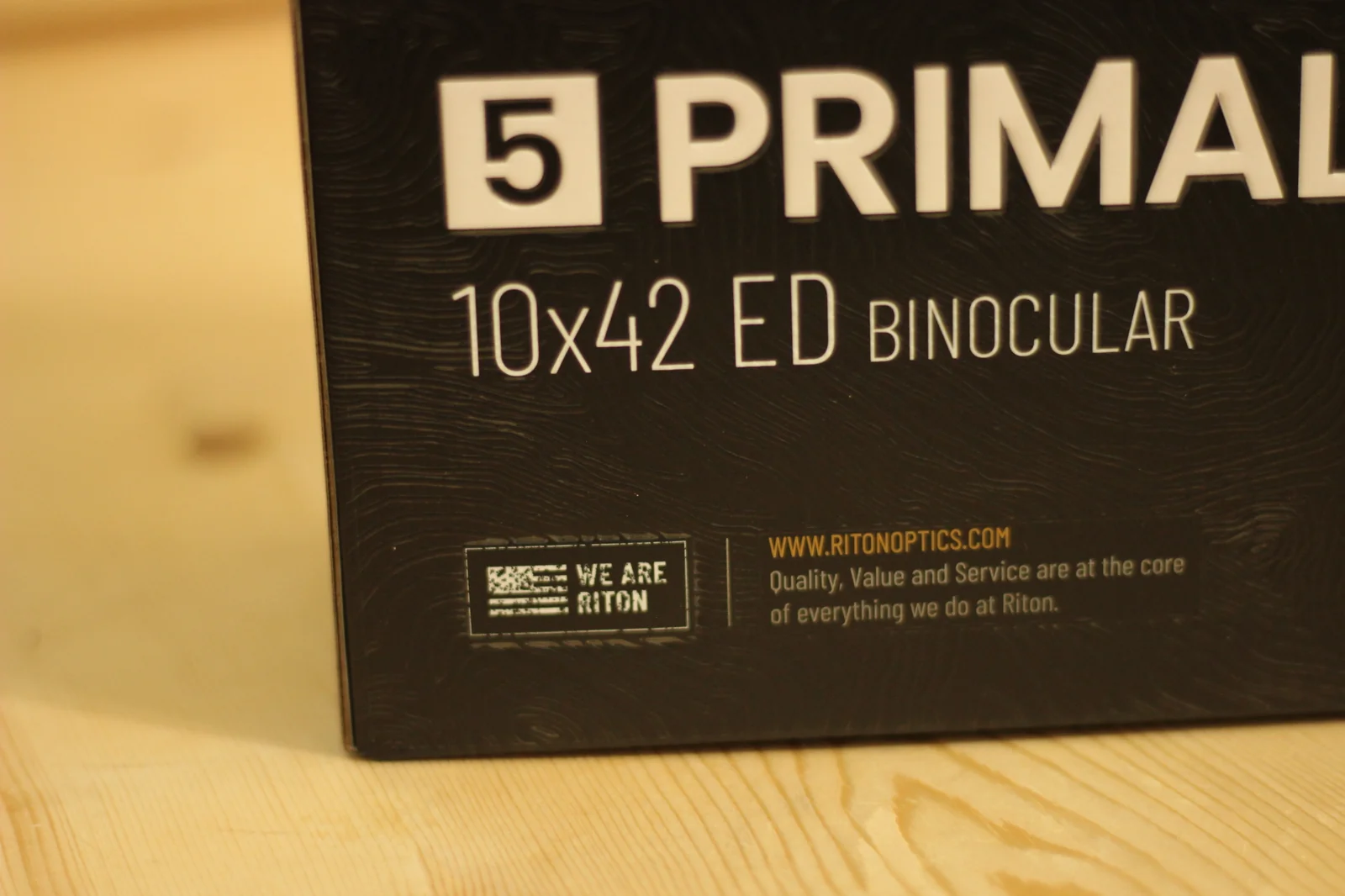Lower section of the Riton 5 Primal 10x42 ED retail box showing the 5 PRIMAL 10x42 ED BINOCULAR wordmark, the WE ARE RITON American-flag badge, and the orange Riton Optics website URL with the brand tagline Quality, Value and Service are at the core of everything we do at Riton