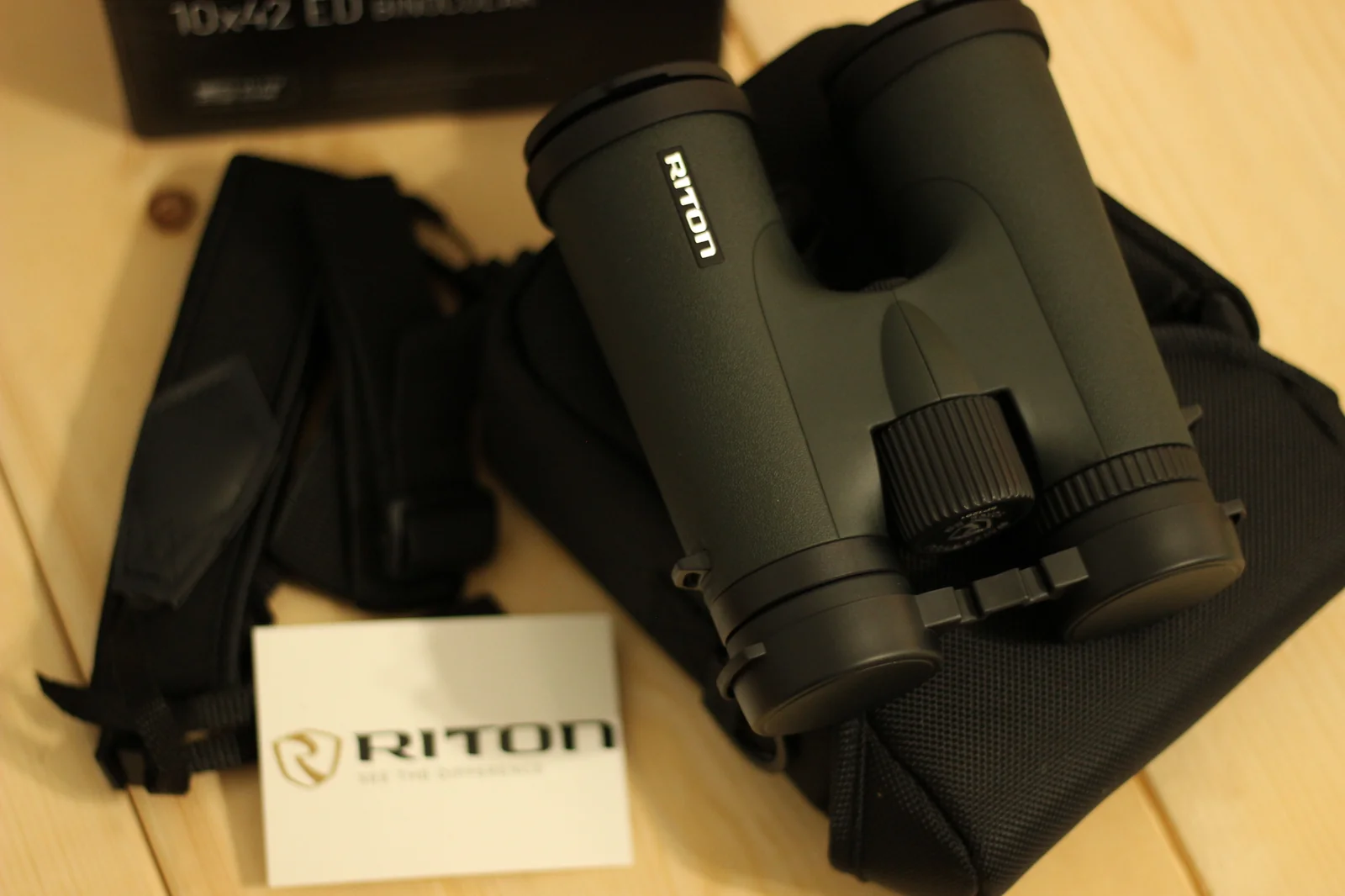 The Riton 5 Primal 10x42 ED unboxed on a pine workbench surface, with the binoculars centered facing right, the rubber-armored objective caps in the closed position, the chest harness laid to the left, the box at upper left, the padded carry bag beneath, and a white Riton registration card with the shield logo in the foreground