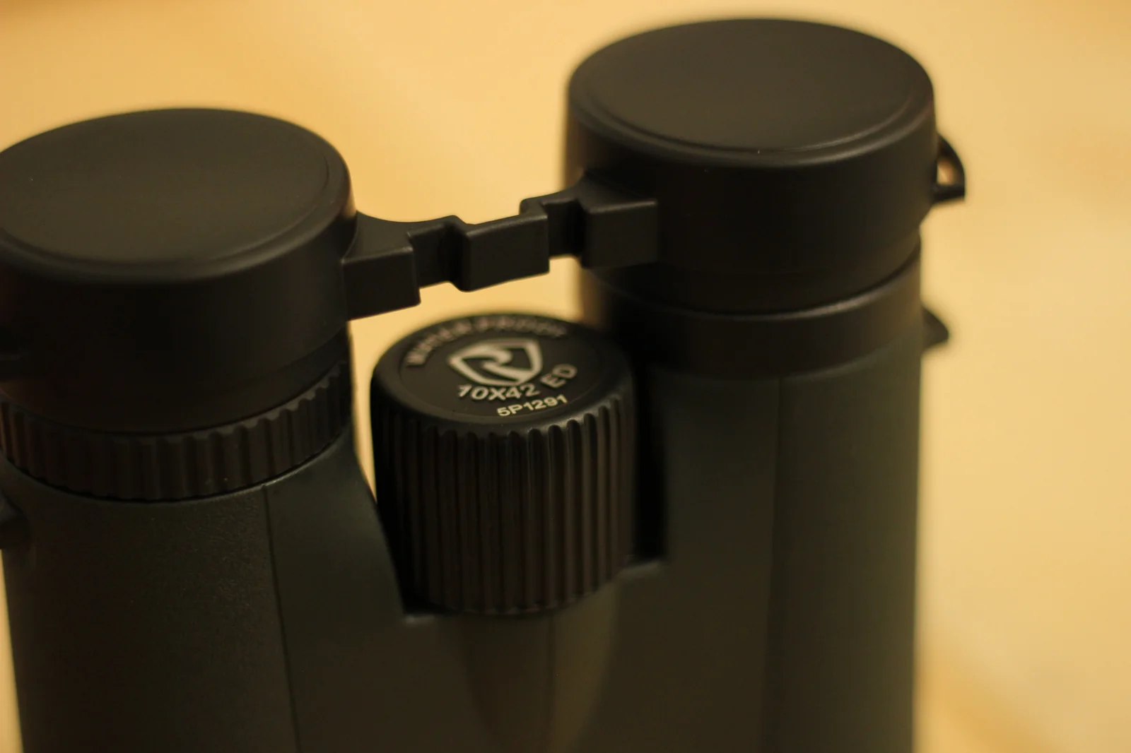 Macro top-down view of the Riton 5 Primal 10x42 ED bridge with the eye cups visible, showing the Riton shield logo on the central diopter cap with WATERPROOF and 10X42 ED model designation, along with the serial number 5P1291 printed below