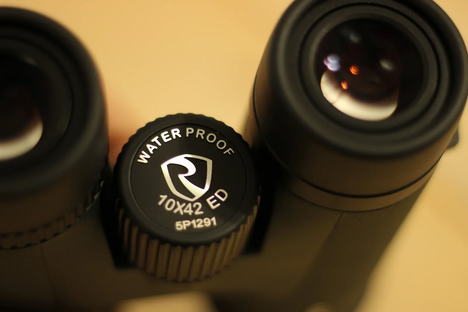 Macro of the Riton 5 Primal 10x42 ED diopter cap on the bridge, showing the WATERPROOF stamping, the Riton shield, the 10X42 ED model designation, and the 5P1291 serial number, flanked by the two eyepieces — the right eyepiece in focus with its inner lens visible