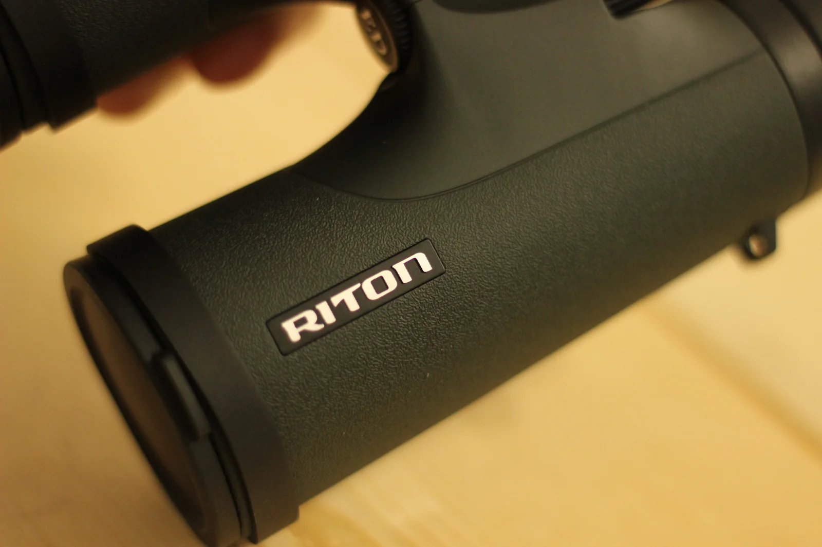 Macro close-up of the front objective lens of the Riton 5 Primal 10x42 ED showing the multi-coated lens surface with a distinct violet and amber tinted reflection from the ED glass coating, rubber armored housing visible around the lens edge