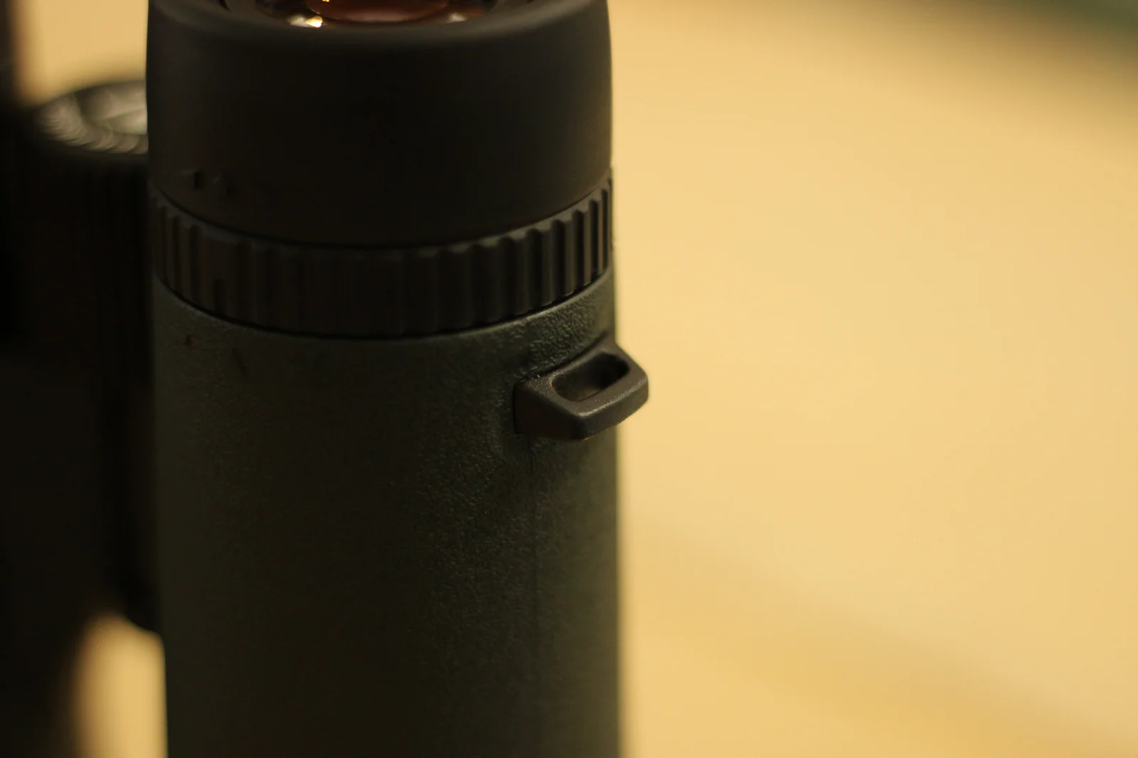 Macro detail of the Riton 5 Primal 10x42 ED focus collar near the eyepiece end of one barrel, the heavily ridged focus collar visible across the top of the frame, with the rubber-armored barrel and the protruding strap lug below it, and the inner objective lens visible at top of frame