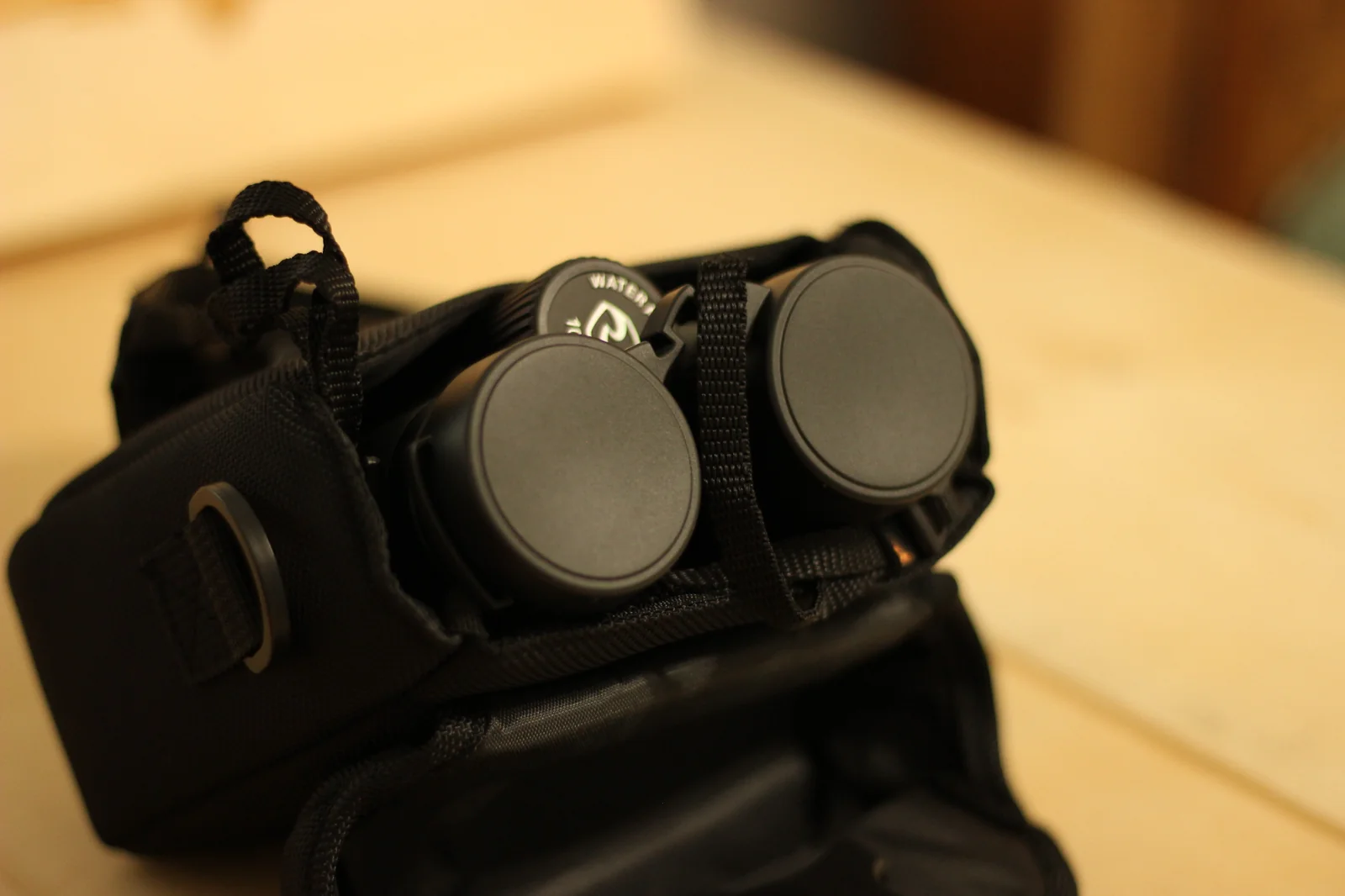 Tight macro of the Riton 5 Primal 10x42 ED focus collar at the eyepiece end of one barrel, with the heavily ridged focus collar at top of frame, the strap lug protruding to the lower right side of the barrel, the rubber-armored barrel taper visible below, and an out-of-focus warm yellow background