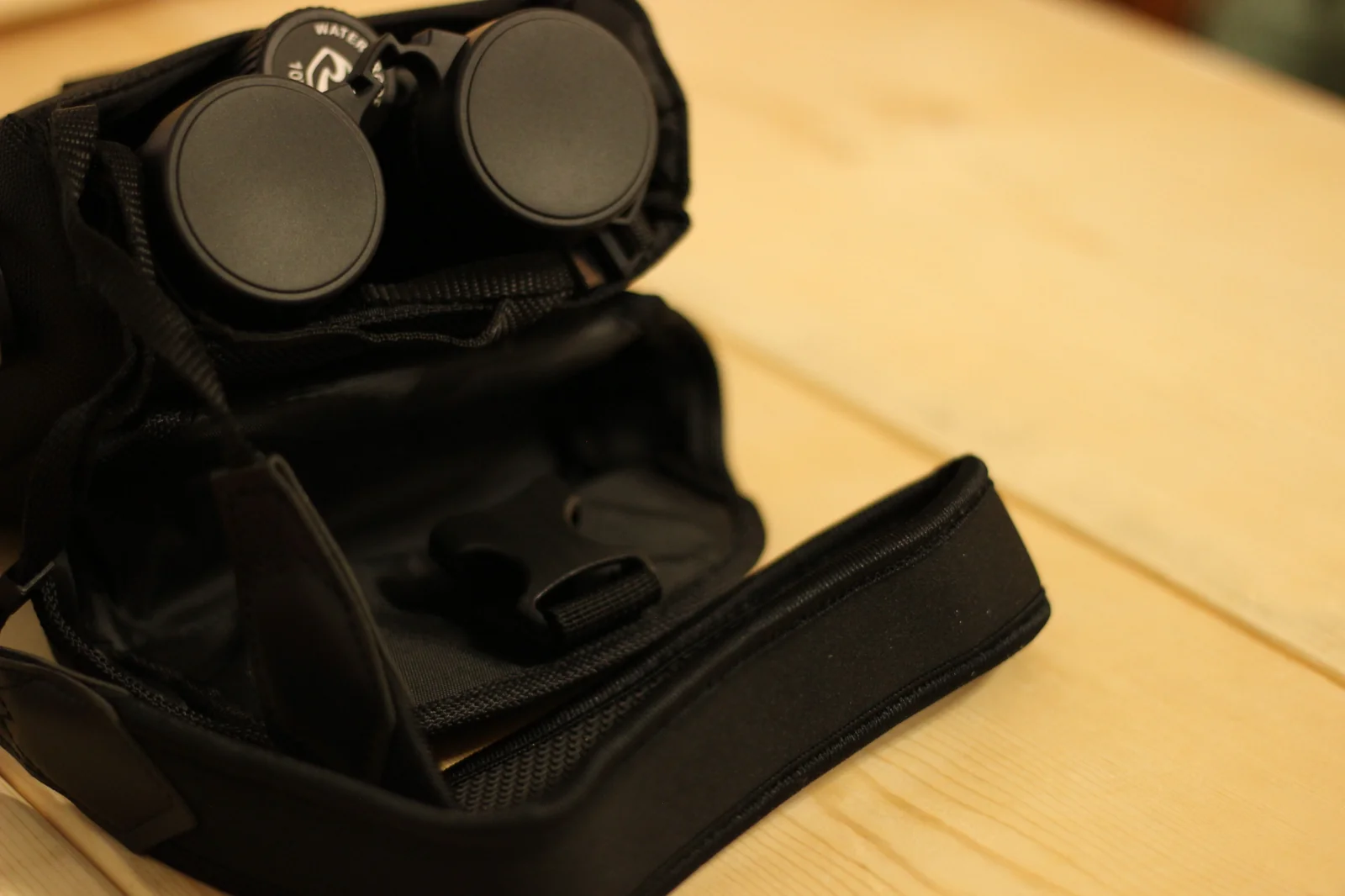 The Riton 5 Primal 10x42 ED seated in the open carrying bag with the objective lens caps in the closed position facing the camera, the WATERPROOF diopter cap on the bridge visible at top, and the contoured chest harness webbing wrapped around the bridge between the two barrels