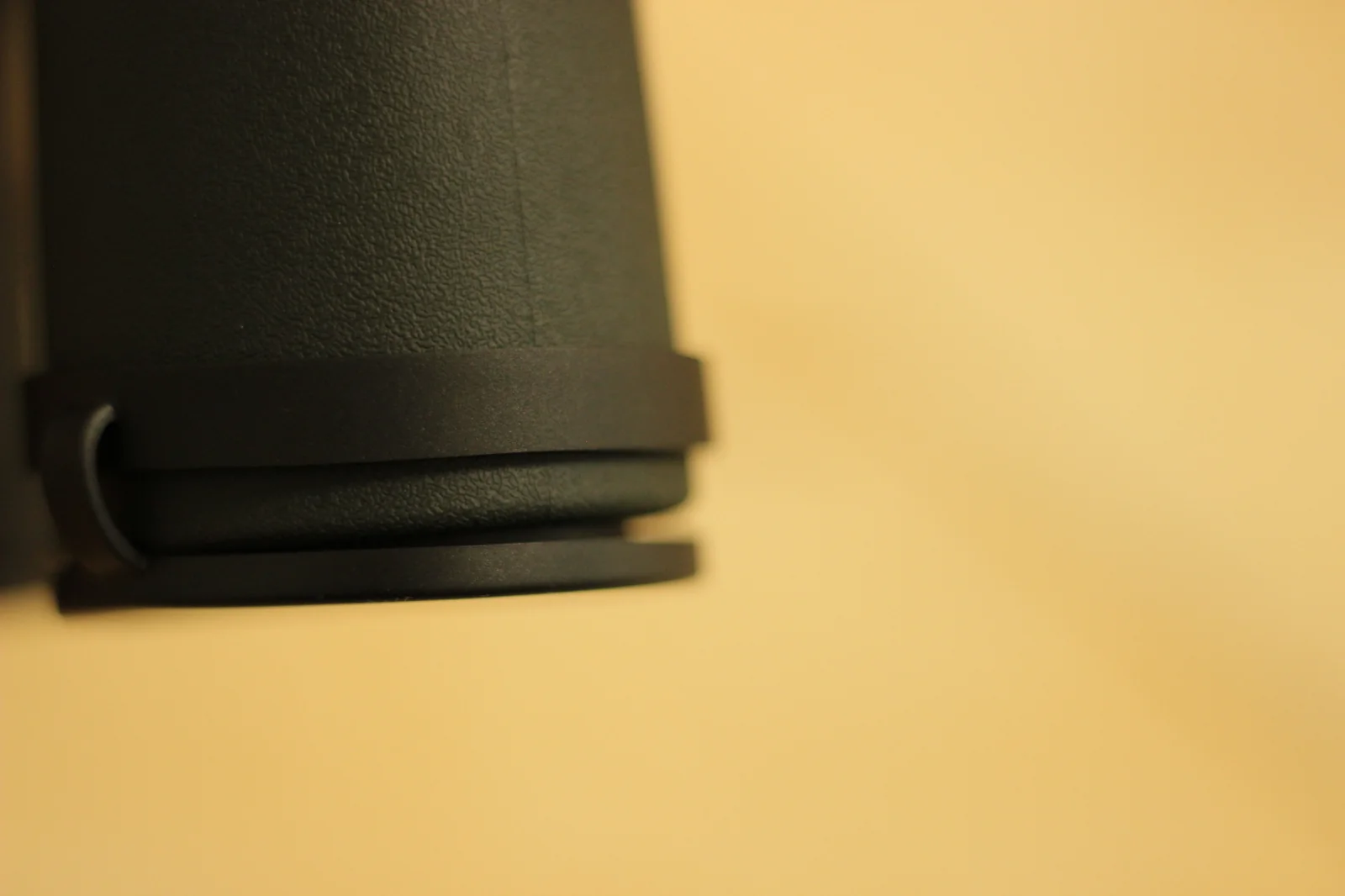 Macro detail of the objective end of one Riton 5 Primal 10x42 ED barrel, showing the rubber-armored barrel taper to the objective bezel, with the strap lug protruding from the lower side of the barrel, photographed against an out-of-focus warm yellow background