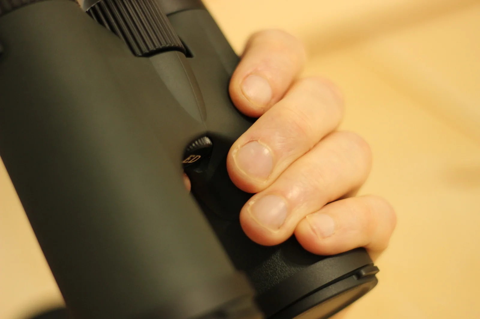 Detail close-up of an adult hand wrapped around one barrel of the Riton 5 Primal 10x42 ED, showing the fingers contacting the rubber-armored housing on the side and underside, with the focus wheel at the top of the frame and the small ED logo medallion at the bottom of the bridge visible between the fingers