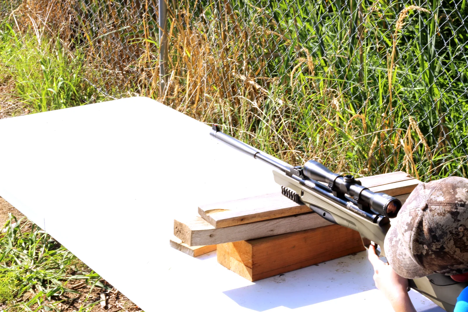 The scoped rifle resting across two blocks of wood on a folding table with the shooter behind it
