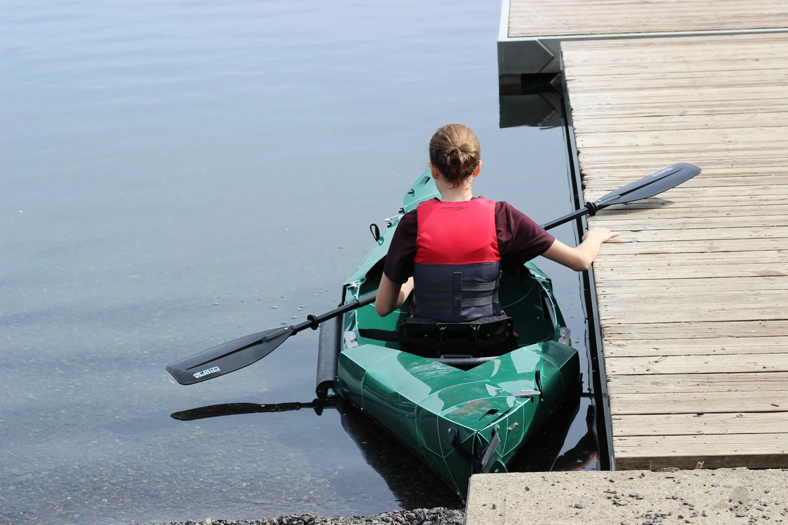 Woman in a red life jacket boarding the green Tucktec Pro kayak from a wooden dock with the paddle in hand