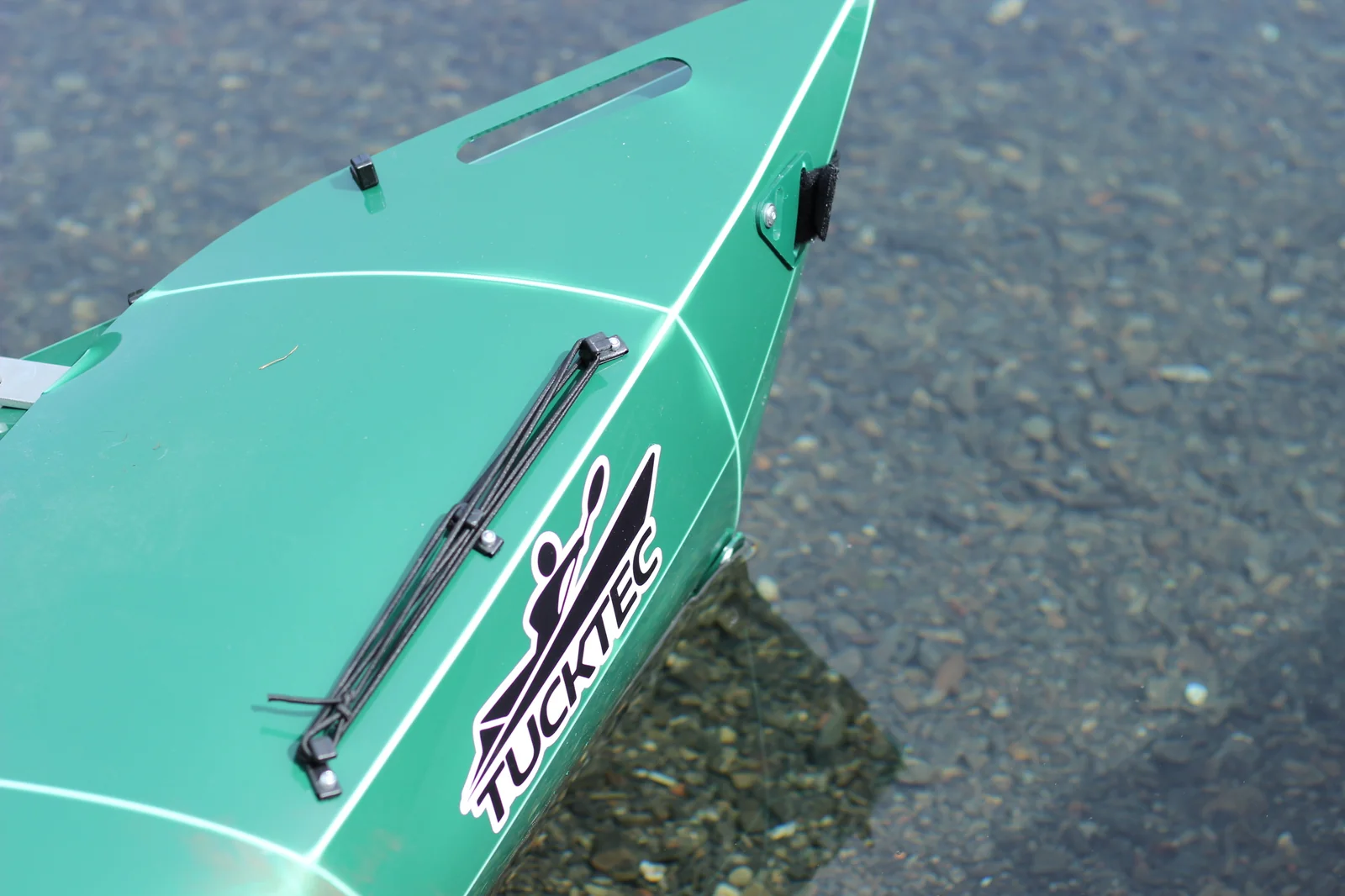Bow of the Tucktec Pro kayak with the TUCKTEC logo and bungee deck rigging visible, sitting on the pebbled shoreline