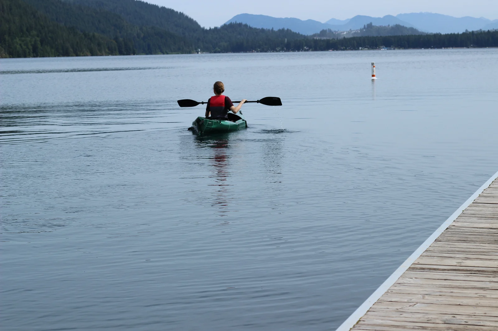 Paddler in a red life jacket paddling the Tucktec Pro kayak away from the dock with mountains and a navigation marker buoy in the distance