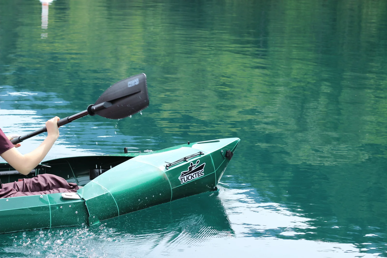 Side view of the Tucktec Pro kayak on glassy reflective green water with the paddle blade lifting out of the water mid-stroke