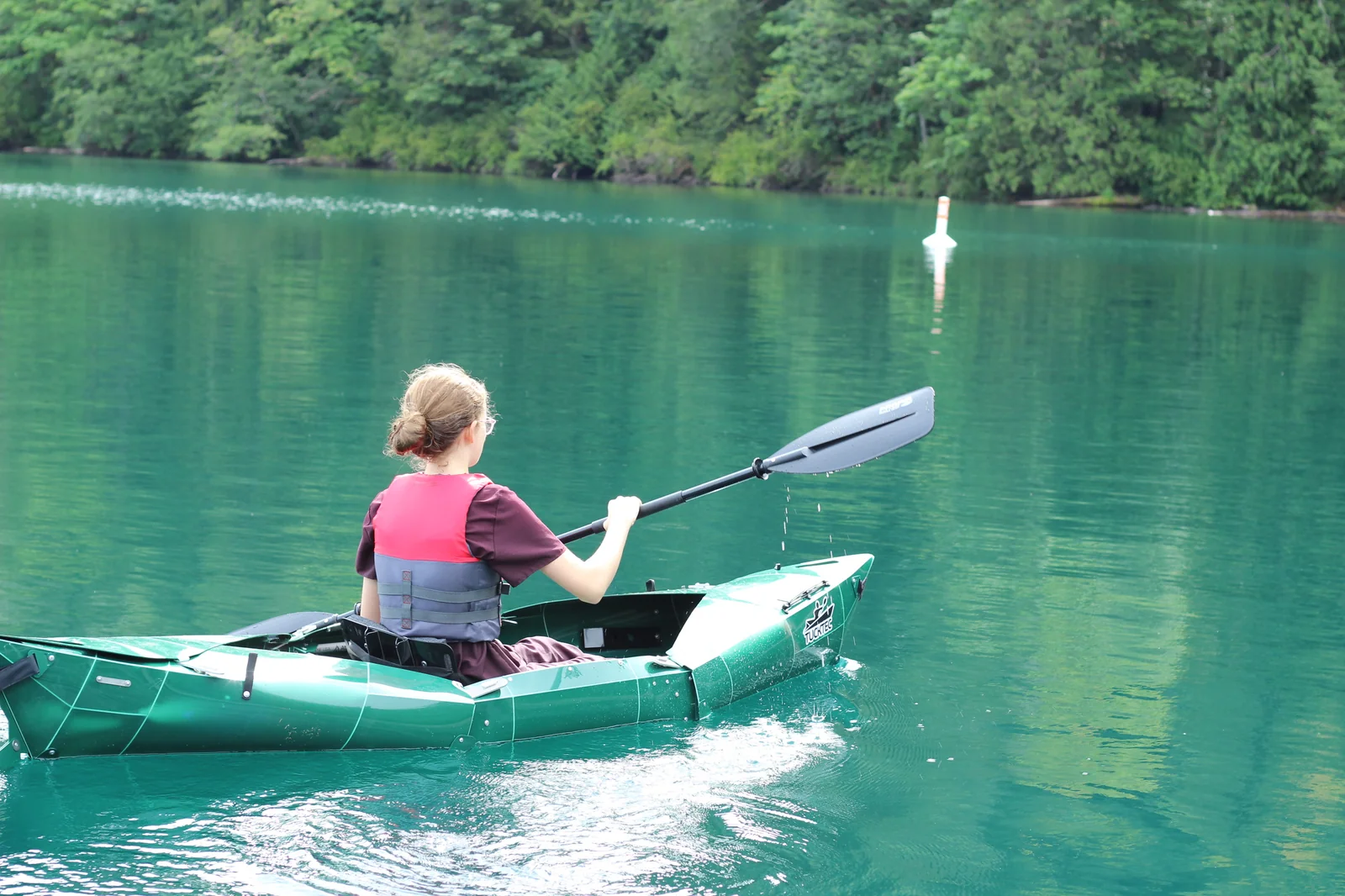 Paddler in a red life jacket on the Tucktec Pro kayak from behind, paddling across green water toward a distant orange navigation marker buoy with forested shoreline beyond