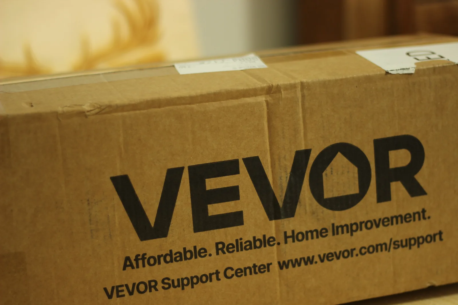 A long brown corrugated VEVOR carton on a pale wooden bench with shipping labels on the top face