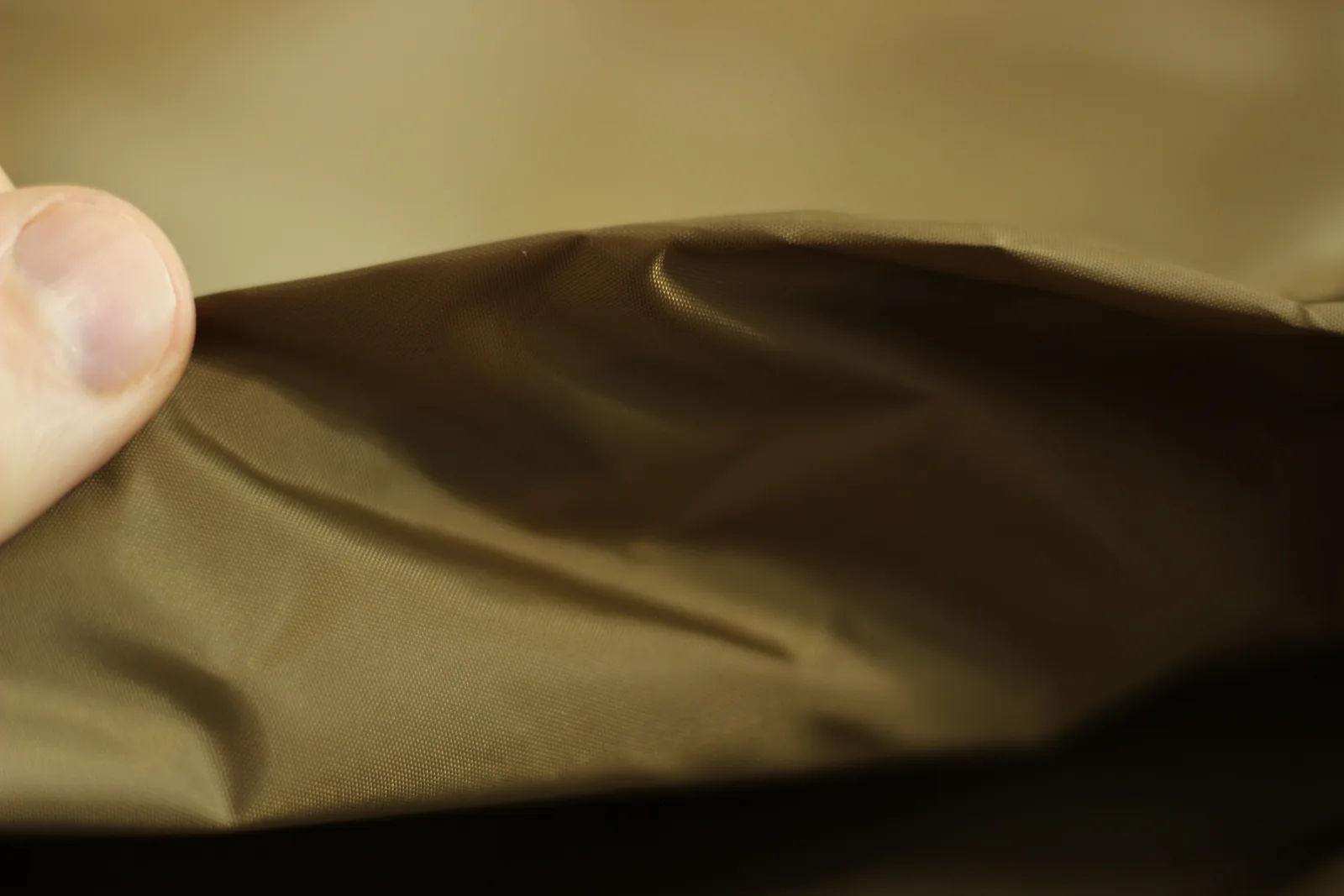 A hand holding up a fold of the coyote-brown canopy fabric showing its fine weave and the sheen of the coating