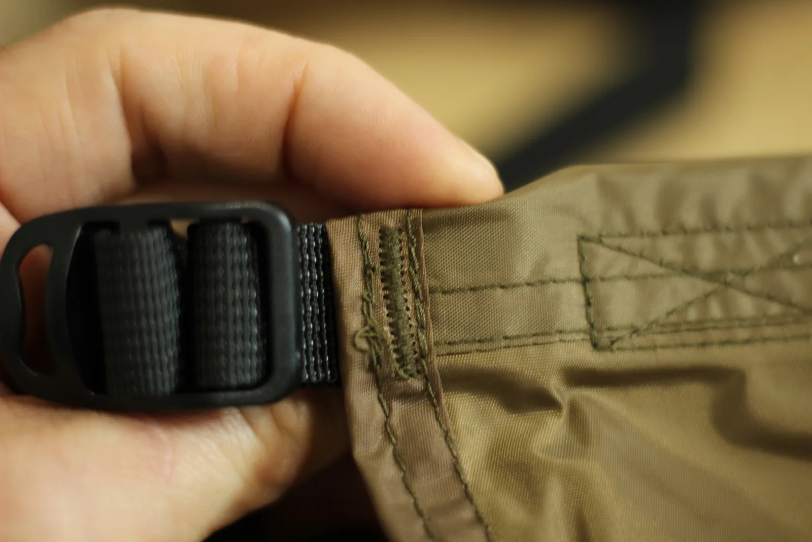 A hand holding a black plastic ladder-lock tensioner threaded with webbing where it joins the tent hem