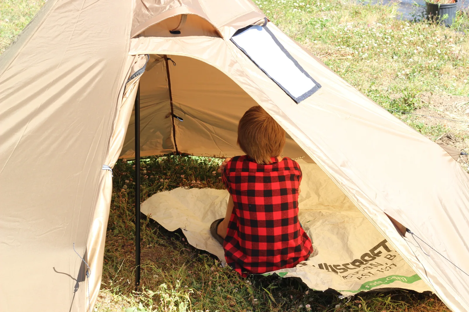 A person in a red plaid shirt sitting inside the pitched tent on a tarp with the door open to the grass outside