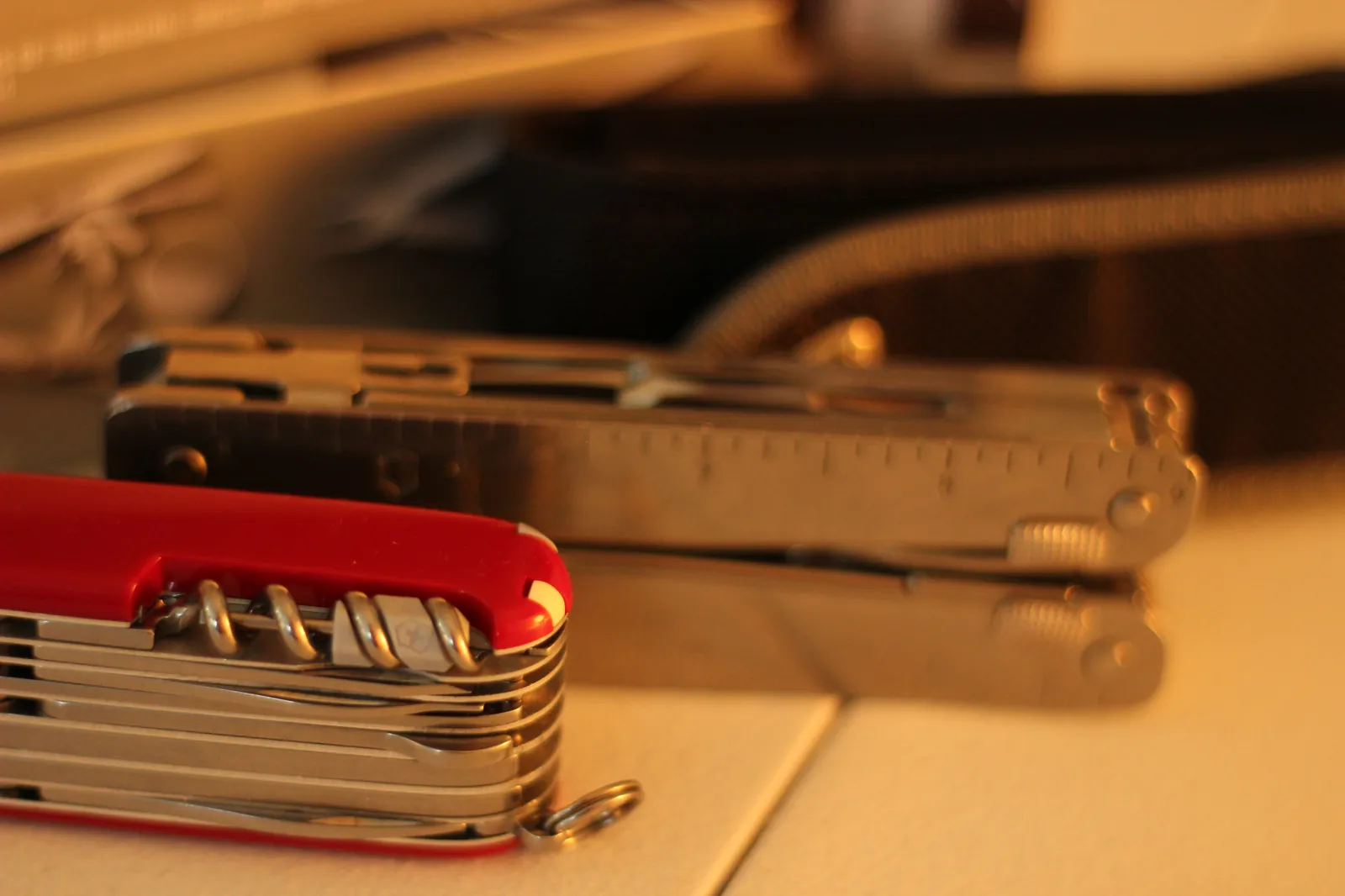 Both products in tight side-by-side close-up — the closed red SwissChamp showing the corkscrew loop in the foreground, the closed Swiss Tool MX Clip with its ruler edge in the background, and the nylon belt pouch beyond