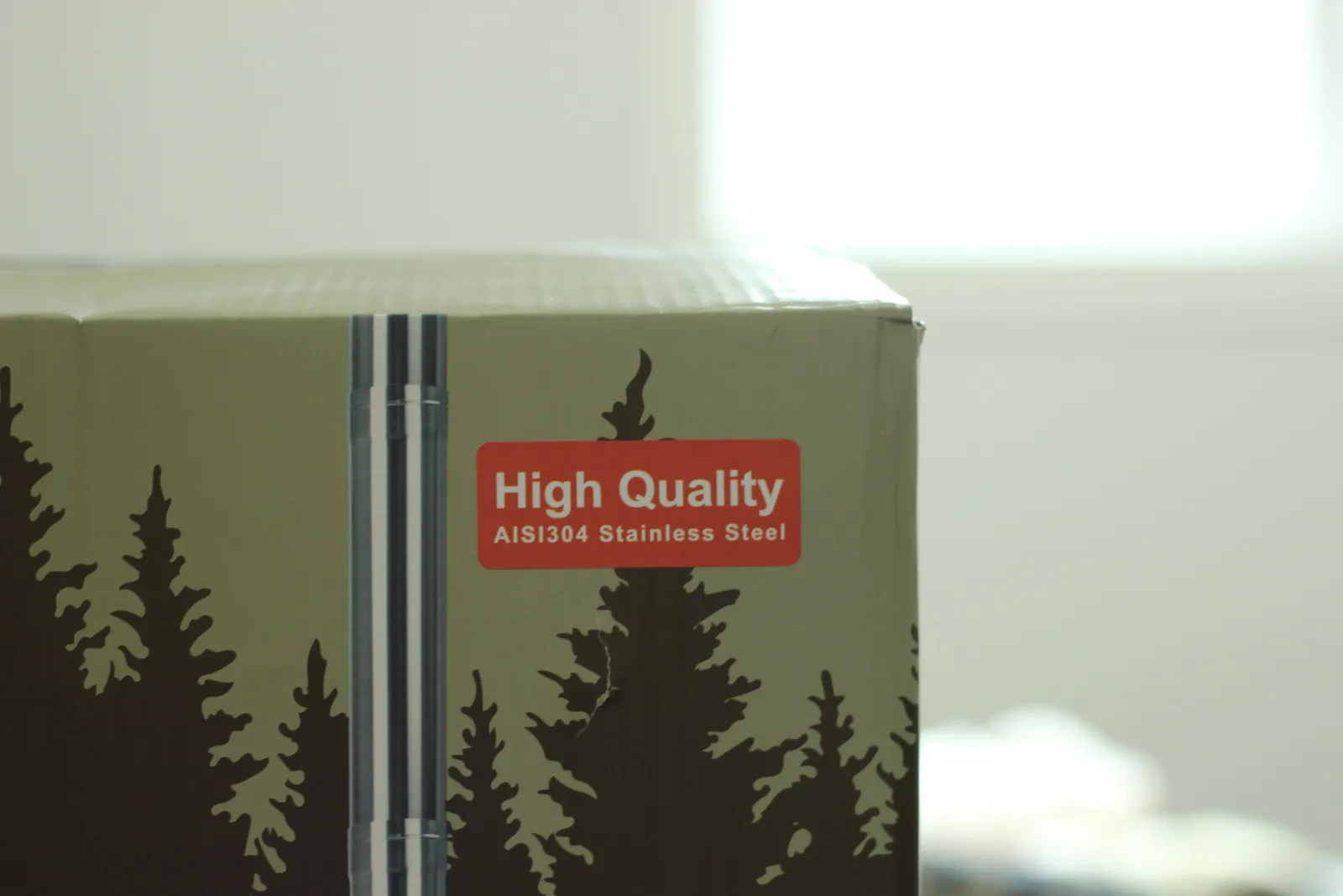 A red sticker on the olive-green carton reading High Quality AISI304 Stainless Steel, over a printed pine forest silhouette