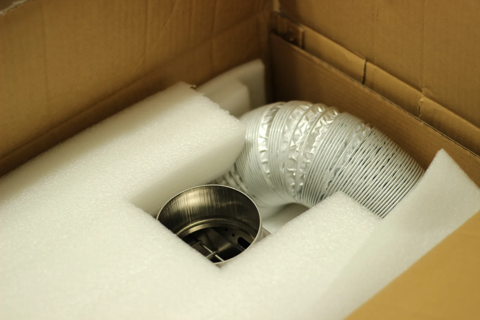 The opened carton showing white foam blocks and a coiled silver flexible external air hose alongside the stainless flue collar