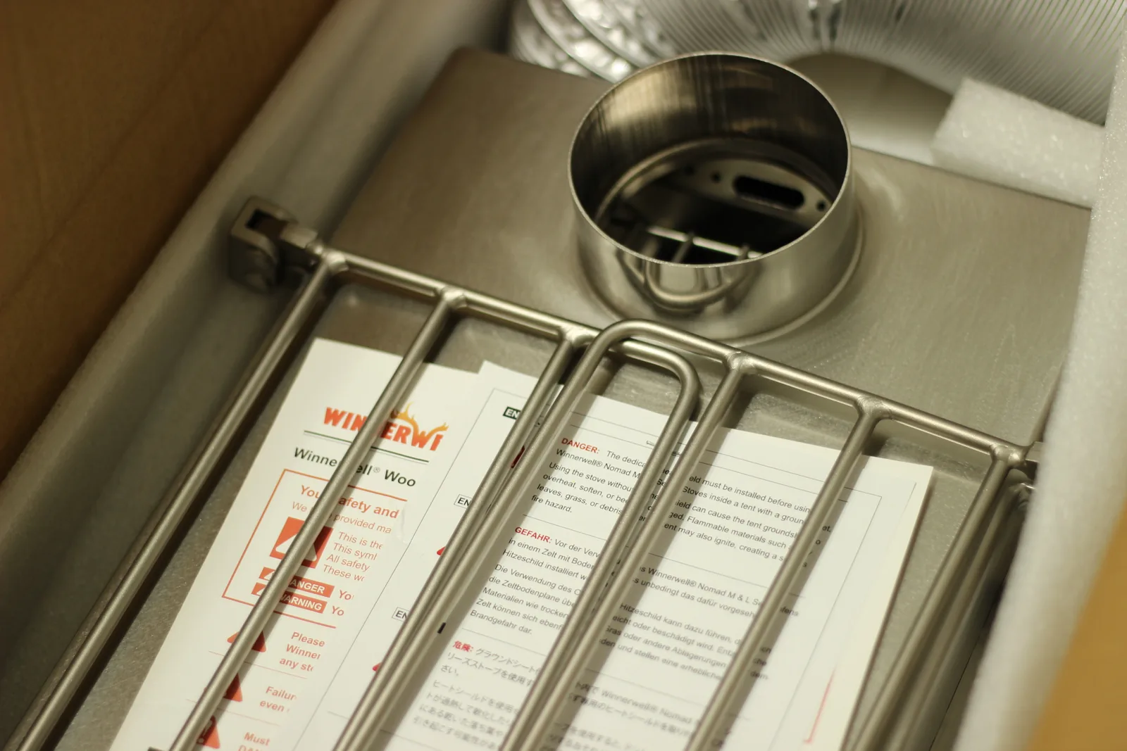 The stove top with its flue collar and wire side rack, sitting over a stack of multilingual printed safety sheets