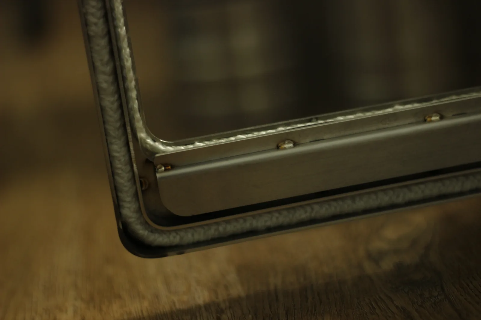 The corner of the stove door showing the glass panel retained by a bolted stainless bar over a white braided ceramic rope gasket
