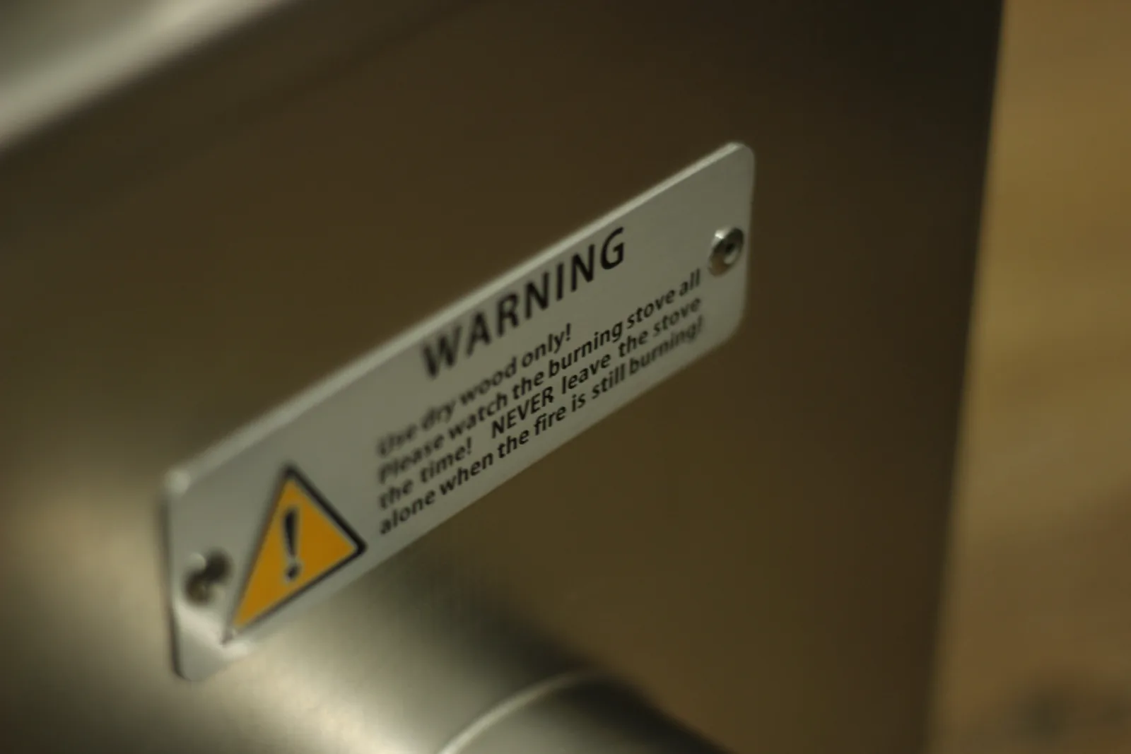 A riveted stainless warning plate on the stove reading Use dry wood only, please watch the burning stove all the time, never leave the stove alone when the fire is still burning