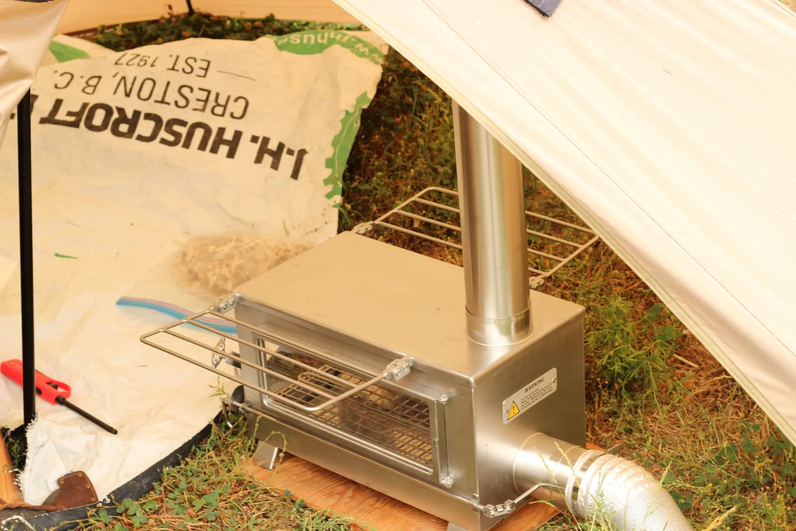 The stove set up inside the coyote-brown tent with its chimney rising through the stove jack and the silver air duct running out under the tent hem