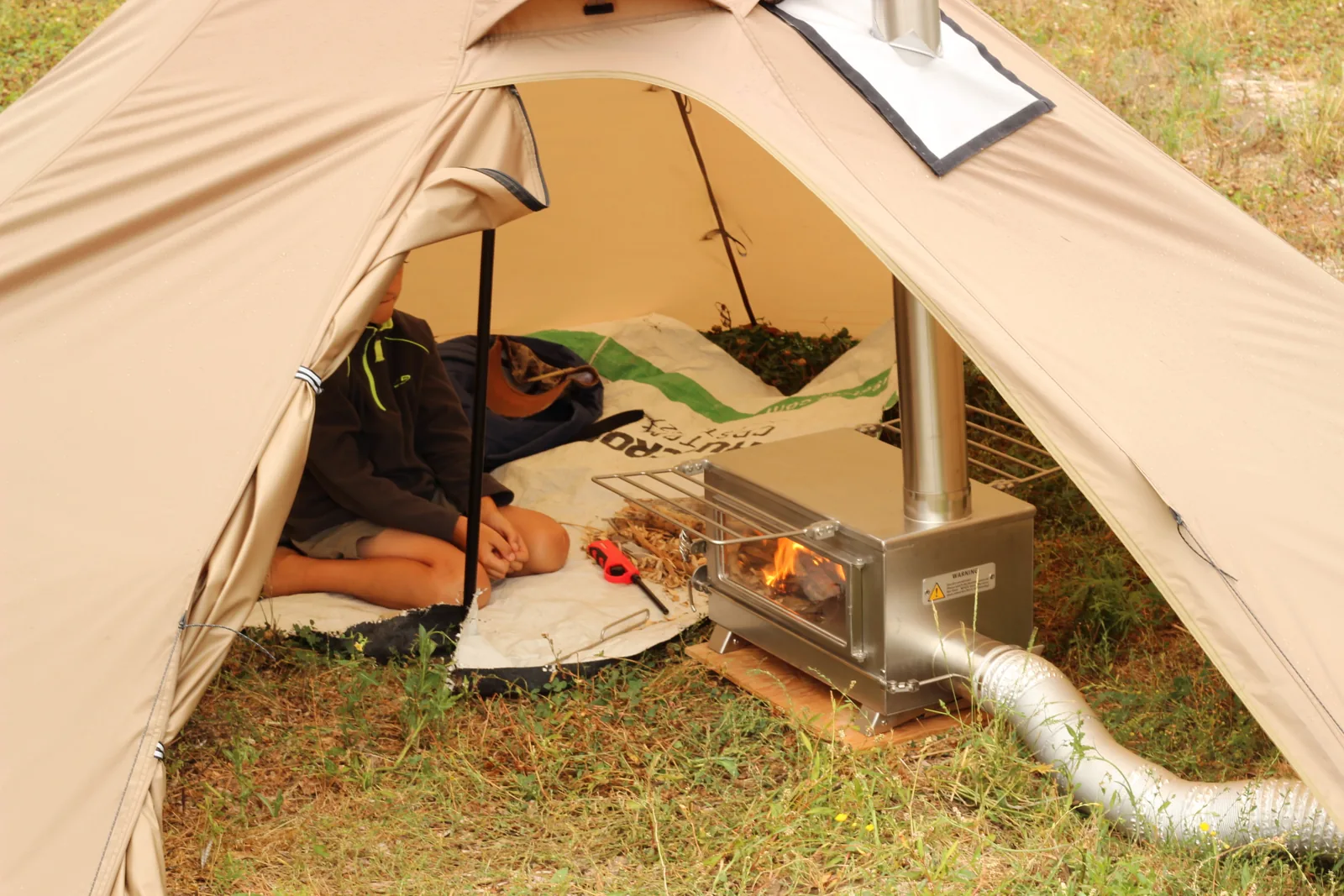 View into the pitched tent with the stove burning just inside the doorway and the external air duct running out across the grass